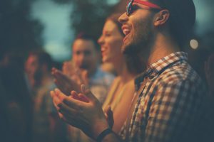 Group of people clapping and cheering after music performance. Standing side by side. There are two guys and one girl and large crowd behind.The closest guy is in focus.Side view.
