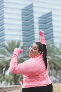 An Hispanic, overweight woman exercising outdoors, lifting handweights. She is looking over her shoulder at the camera, smiling. In the background is a modern office building and palm trees.